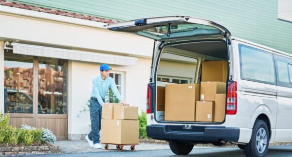 トヨタのピクシス バンをイメージしたシルバーの軽商用バン。開いた後部荷室に段ボール箱を積み込む日本人配送スタッフの様子。軽バンの積載性と仕事での使い方を紹介。
